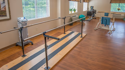 Rehabilitation-style exercise room with parallel walking bars, fitness machines, and wood flooring under large windows.