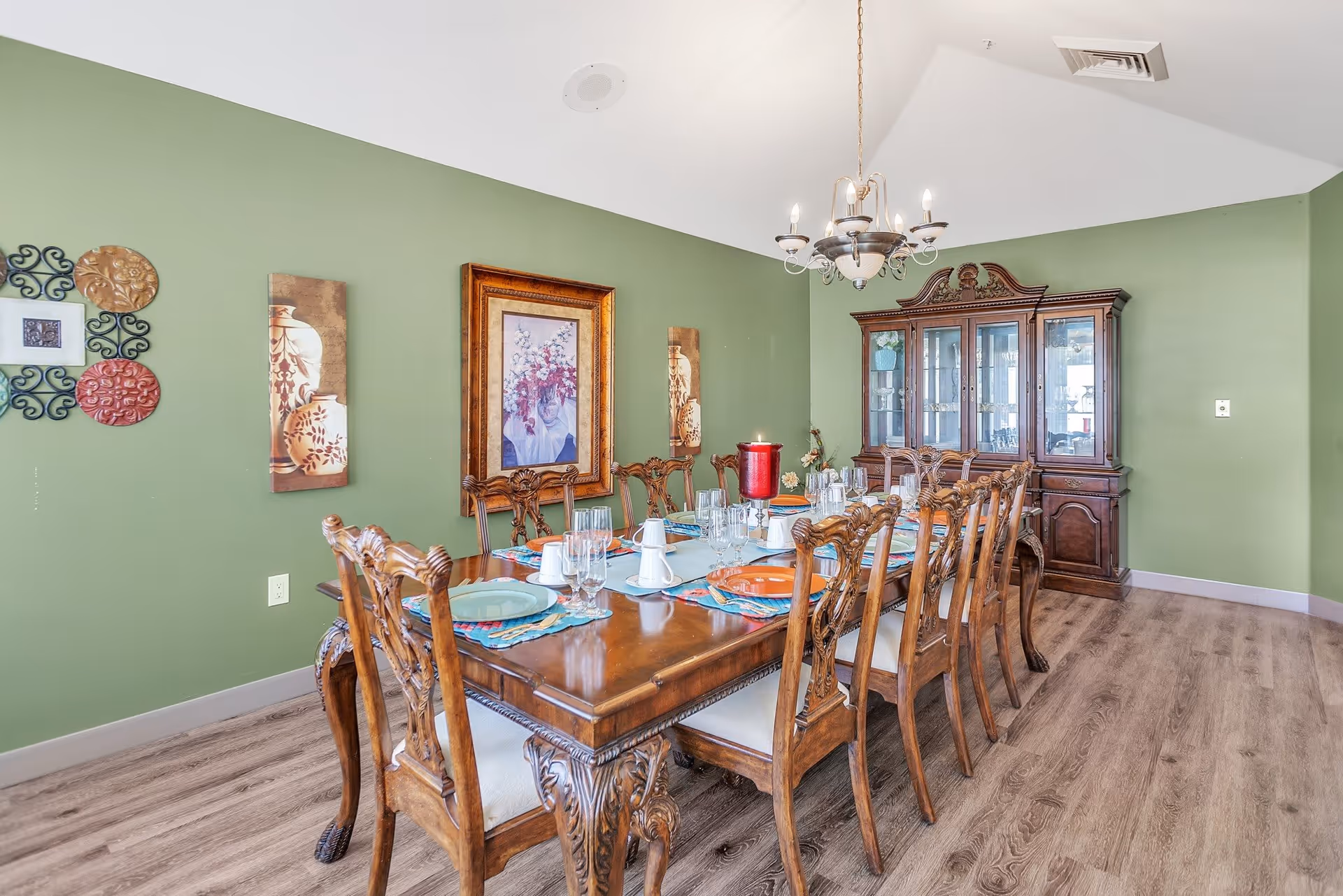 A formal dining room with a long wooden dining table set for ten people. The table is adorned with plates, glasses, and a red candle centerpiece. The room has green walls decorated with framed artwork and a wooden china cabinet. A chandelier hangs above the table, and the floor is covered with wood-style flooring.