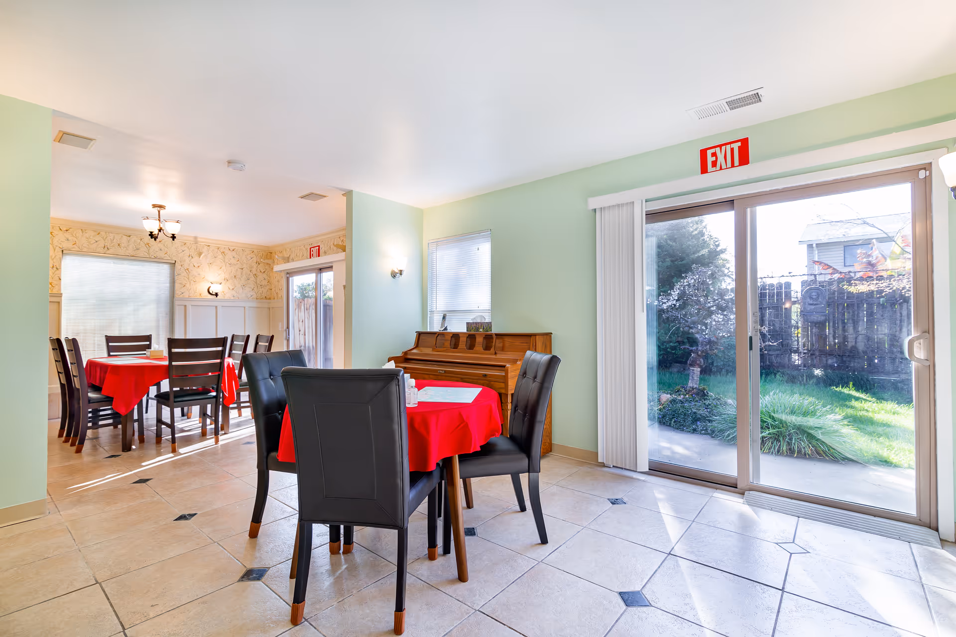 A dining area with tables covered in red cloth, a piano, and a view of the outdoors through sliding glass doors.