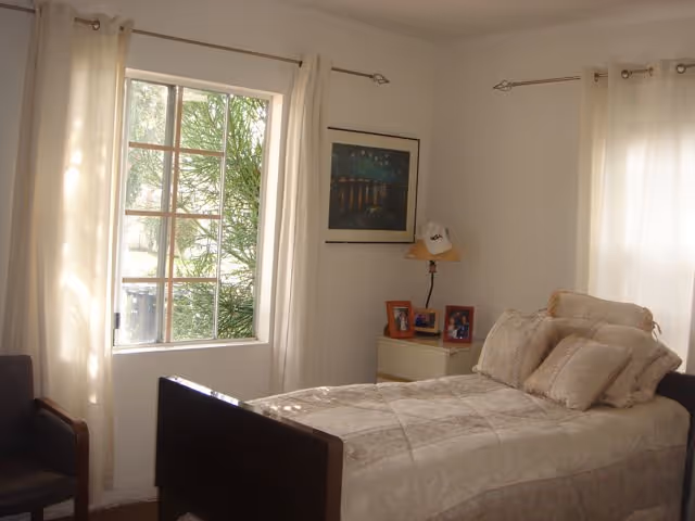 A bedroom with a single bed covered in beige bedding and multiple pillows. There is a window with white curtains letting in natural light, a small nightstand with framed photos and a lamp, a framed picture on the wall, and a dark chair in the corner.