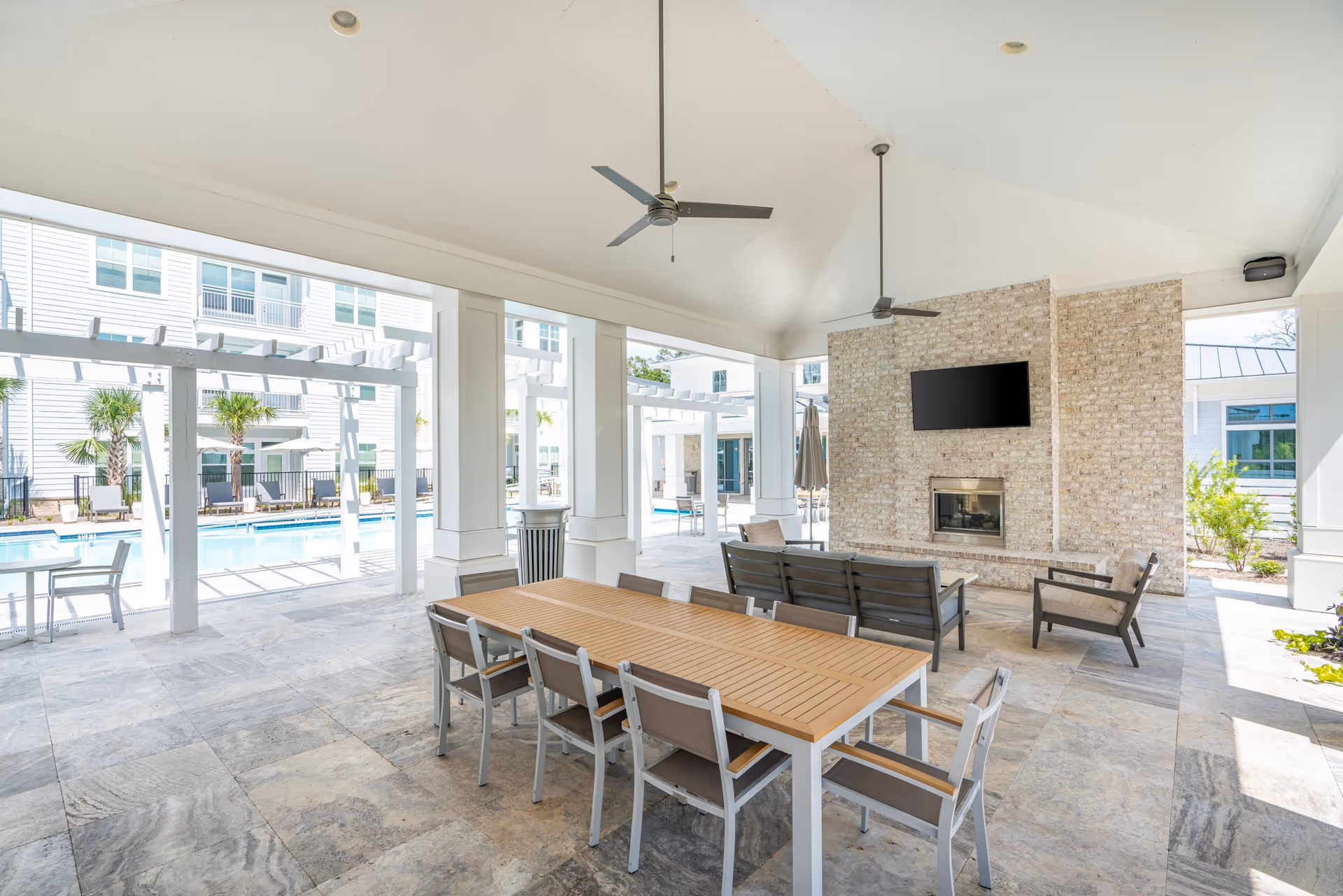 Covered outdoor seating area with a long wooden table and chairs, ceiling fans, a brick fireplace with a mounted TV, and a view of a swimming pool and white pergolas in the background.