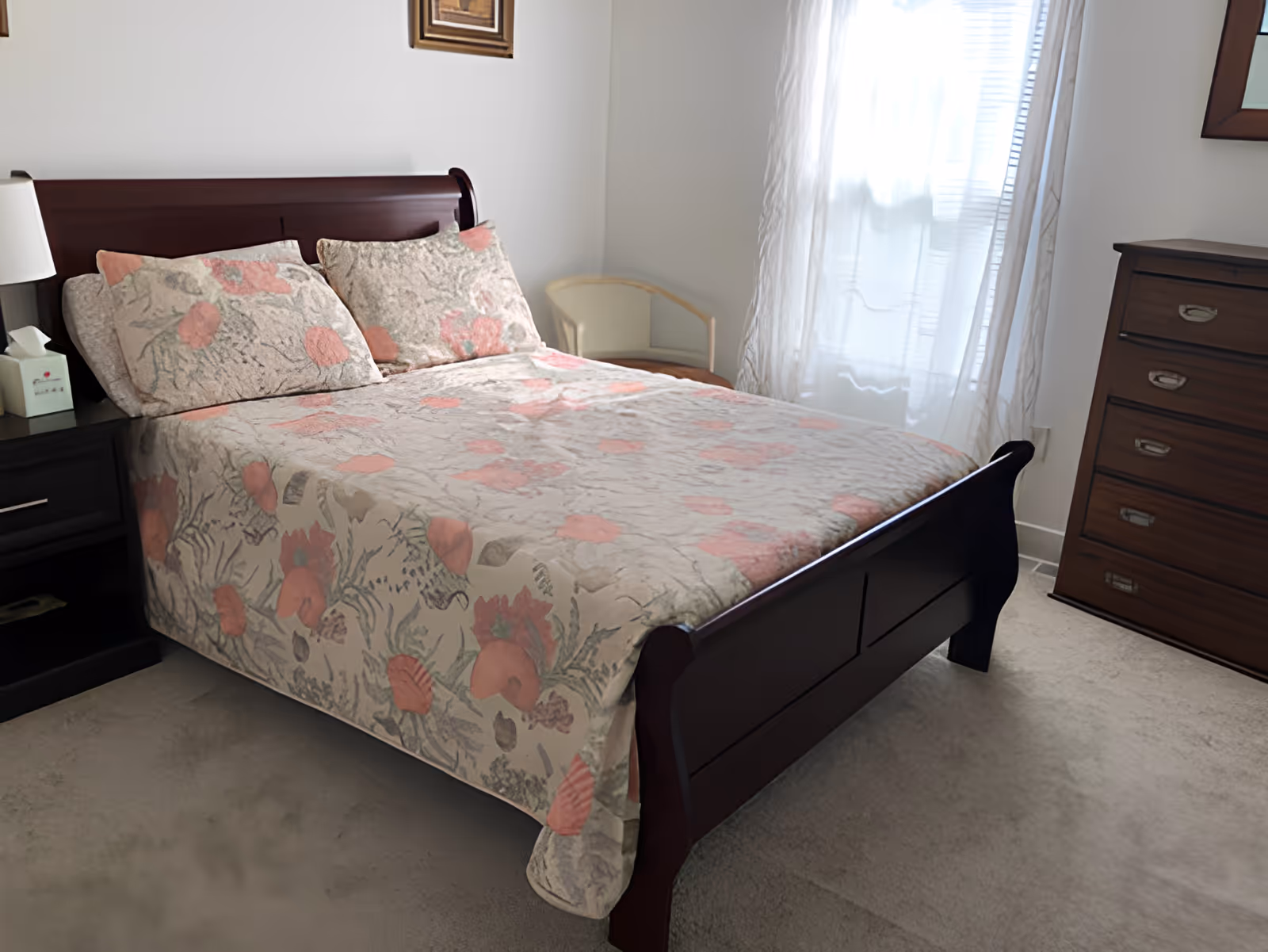Sunlit bedroom with a wooden sleigh bed covered in floral bedding, a nightstand, and a dresser.