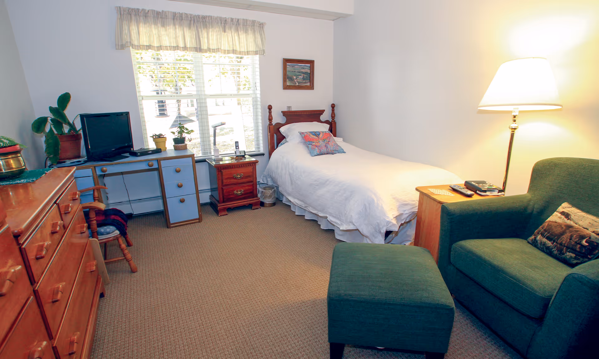 A small bedroom with a single bed covered in white bedding and a colorful pillow. Next to the bed is a wooden nightstand with a lamp and a cordless phone. A green armchair with a matching ottoman is positioned near the bed, accompanied by a floor lamp. Across from the bed is a wooden dresser and a desk with a small television, plants, and a chair. A window with blinds and a valance lets in natural light.