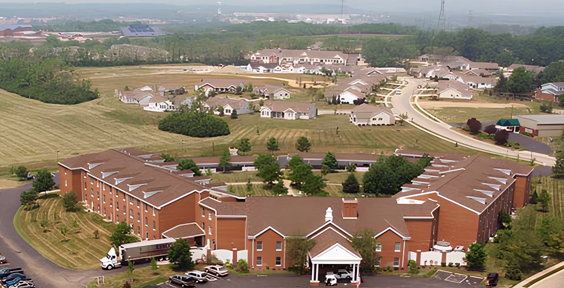 Aerial view of Ohio Living Mount Pleasant senior living facility showing a large, U-shaped main building with a brown roof and red brick exterior surrounded by green lawns, trees, and smaller residential buildings in the background under a clear sky.