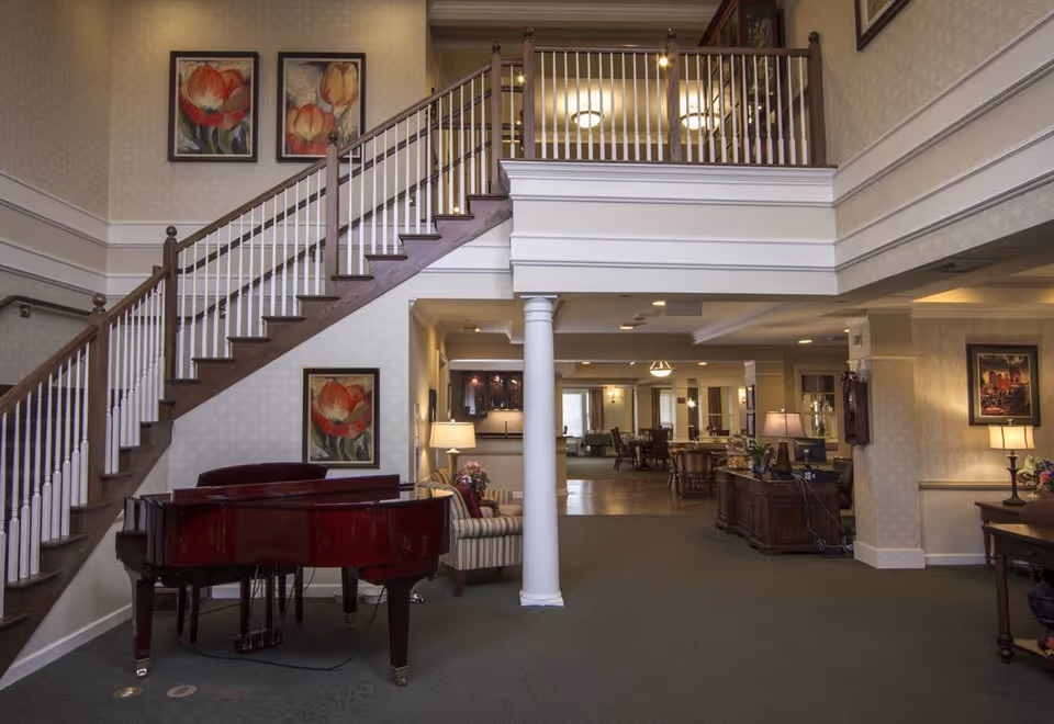Spacious senior living lobby with a grand piano, staircase, seating areas, and a reception desk.