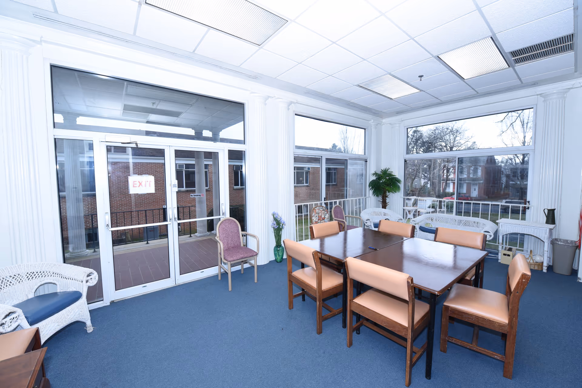 Bright communal dining/activity room with a large wooden table and chairs, wicker seating, potted plants, and large windows overlooking the outside.