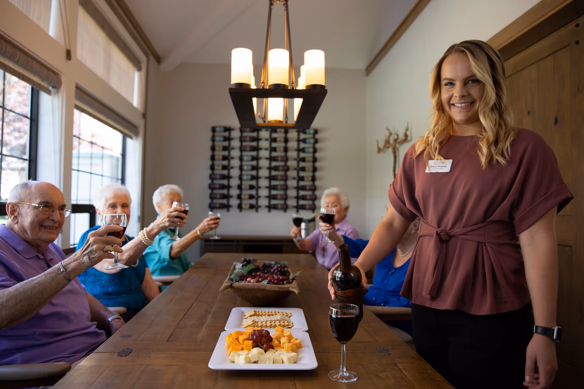 A group of elderly people sitting around a wooden dining table raising glasses of red wine in a toast. A smiling woman stands at the end of the table pouring wine into a glass. The table has plates with cheese, crackers, and grapes. The room has large windows and a wine rack on the wall.
