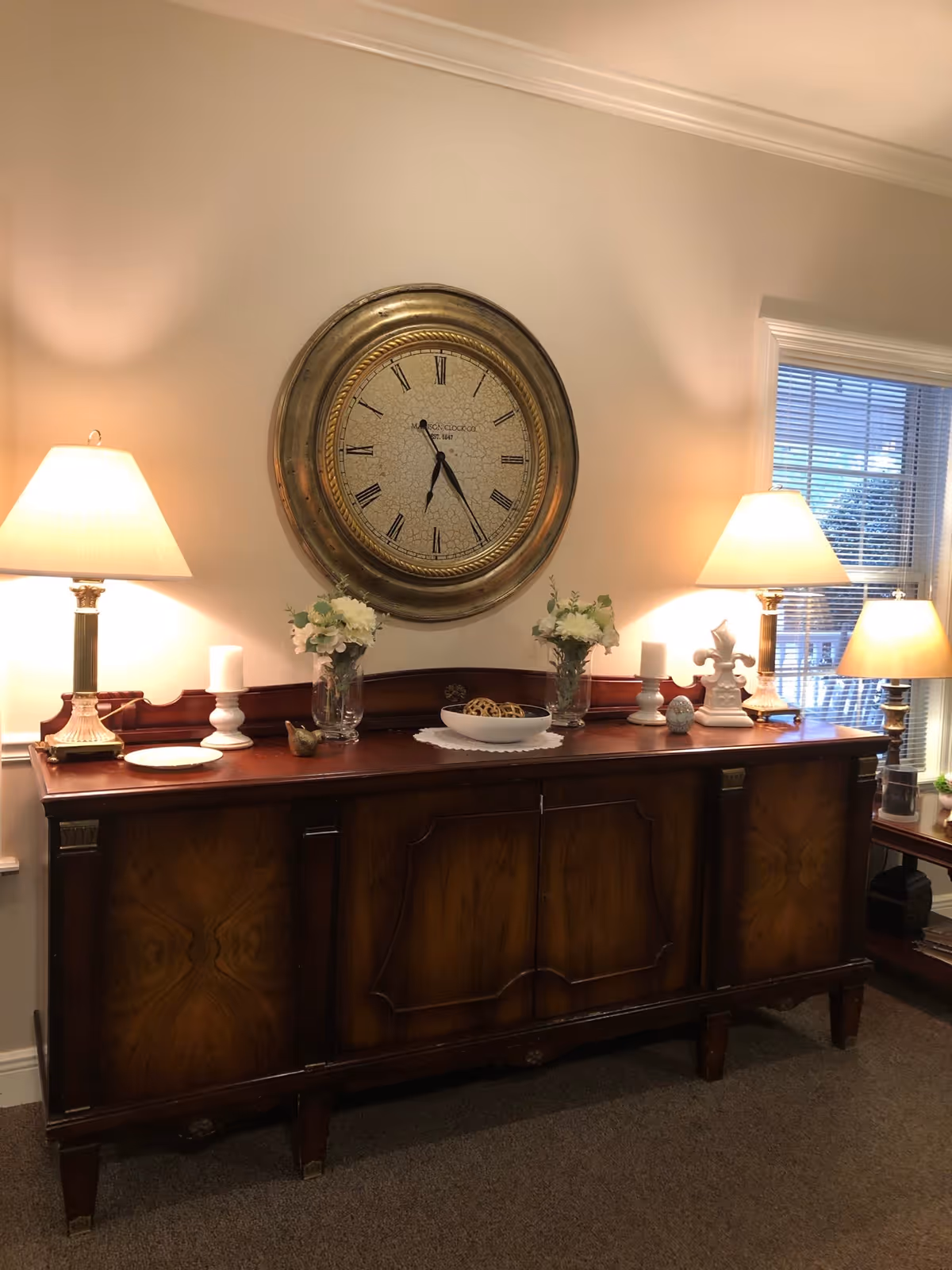 A wooden sideboard with decorative items including two table lamps, two vases with white flowers, candles, and a bowl with decorative spheres. Above the sideboard is a large round wall clock with Roman numerals. To the right, there is a window with blinds partially open, allowing some natural light into the room.
