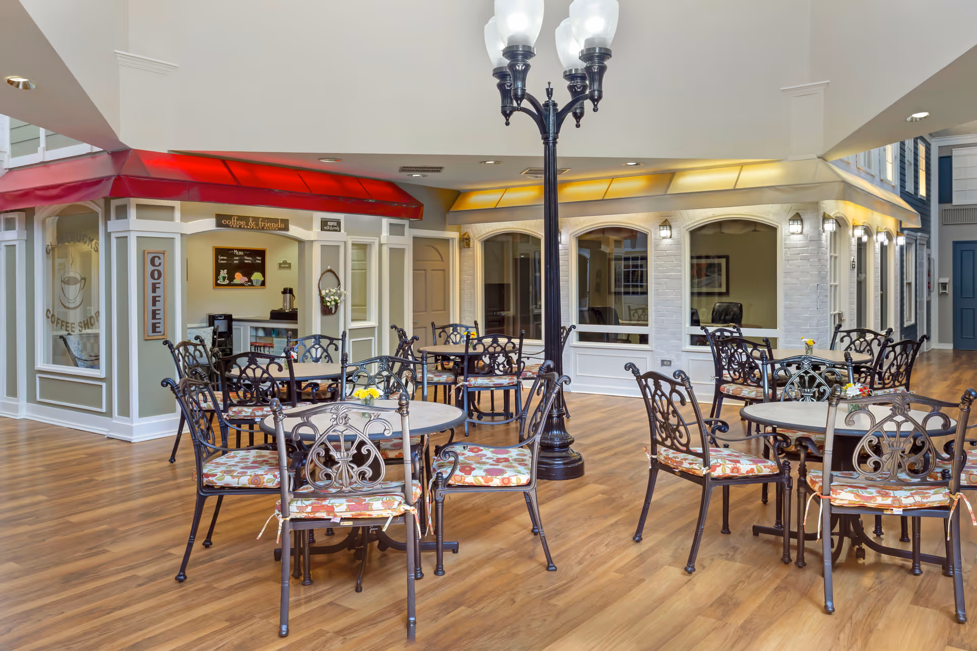 Indoor seating area with multiple round tables and ornate metal chairs with floral cushions, situated on a wooden floor. There is a coffee shop with a red awning labeled 'coffee & friends' and 'COFFEE' on the side. A black streetlamp-style light fixture stands in the middle of the seating area. The surrounding walls have large windows and doors, giving the space a cozy, café-like atmosphere.