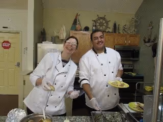 Two chefs in white uniforms standing behind a serving counter in a kitchen, smiling and holding plates of food. The kitchen has wooden cabinets, a microwave, and various kitchen utensils visible in the background.
