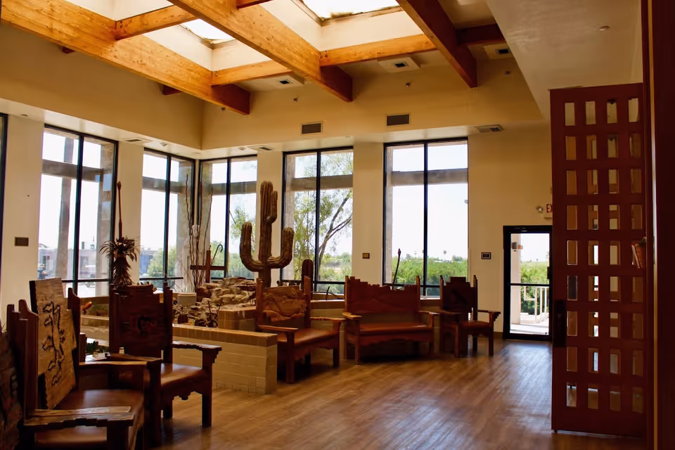 A spacious interior common area with large windows letting in natural light. The room features wooden furniture with carved designs, including chairs and benches arranged around a central tiled planter containing tall decorative cacti. The ceiling has exposed wooden beams and skylights, and there is a door leading outside on the right side.
