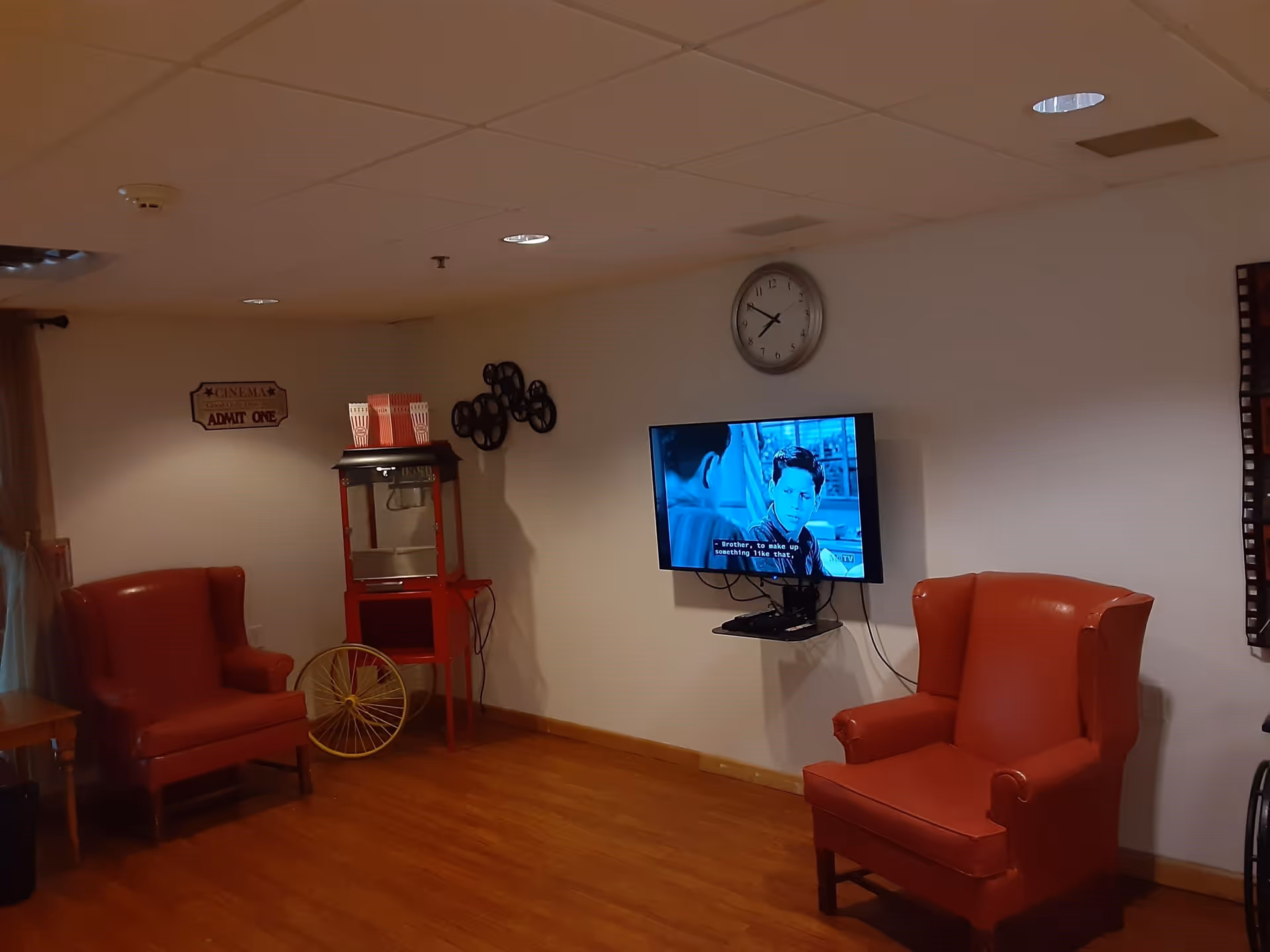 A cozy room with two red armchairs facing a wall-mounted TV showing a black and white movie with subtitles. There is a vintage popcorn machine with popcorn boxes on top, a wall clock above the TV, and decorative film reels on the wall. The floor is wooden, and the walls are light-colored.