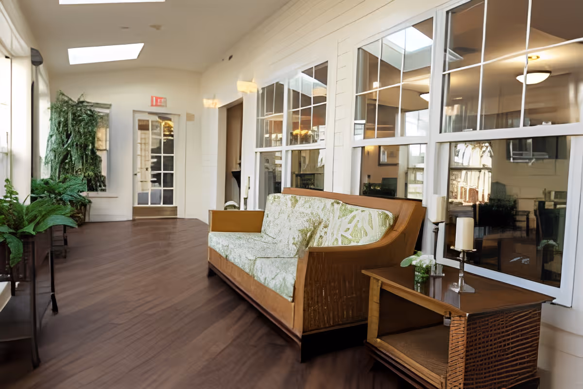 A bright indoor hallway with wooden flooring and large windows on the right side. There is a wooden sofa with green patterned cushions and a matching wooden side table with candles and a small plant. On the left side, there are several green plants placed on stands. The hallway leads to a glass door at the far end with an exit sign above it.