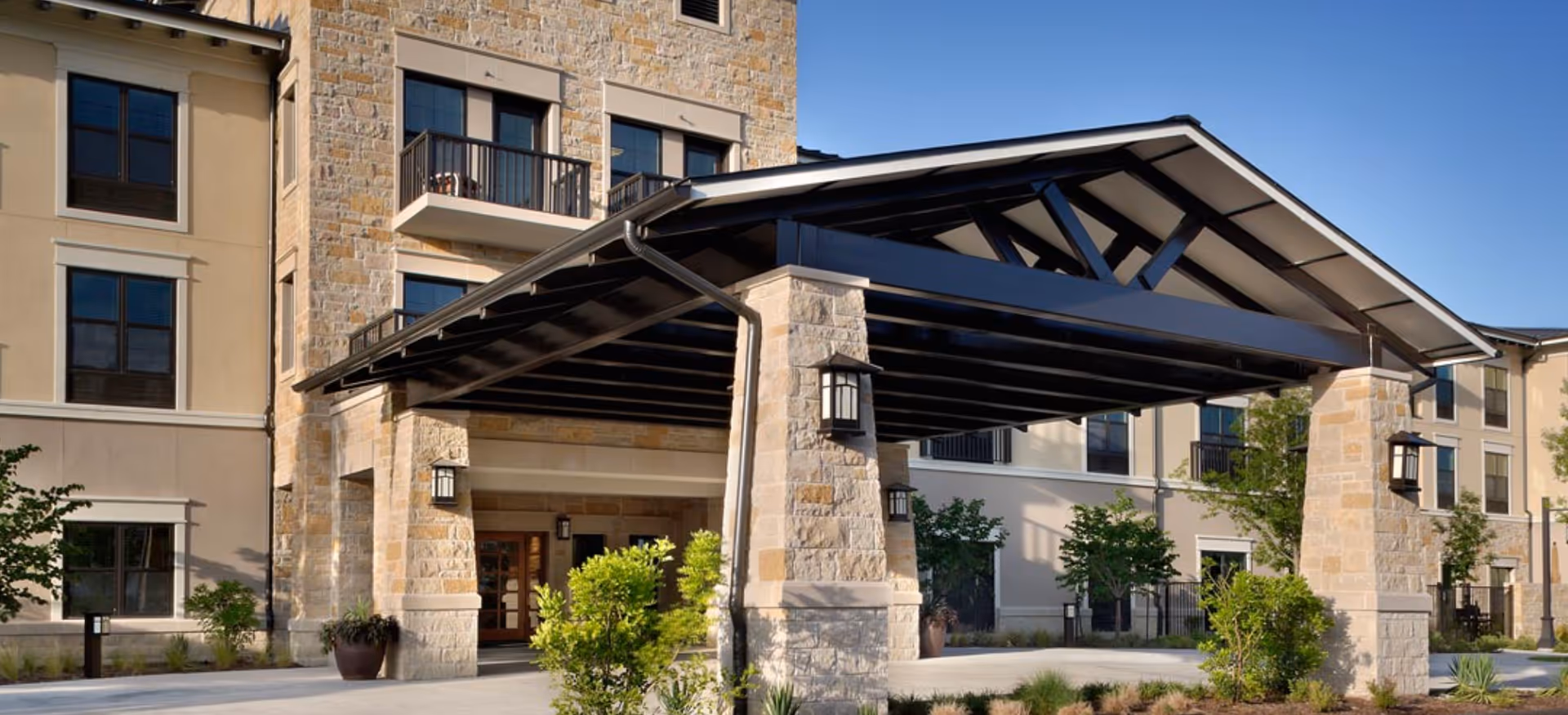 Exterior view of Belmont Village Senior Living West Lake Hills showing the main entrance with a large covered driveway supported by stone pillars, surrounded by landscaping and a multi-story building with balconies and windows.