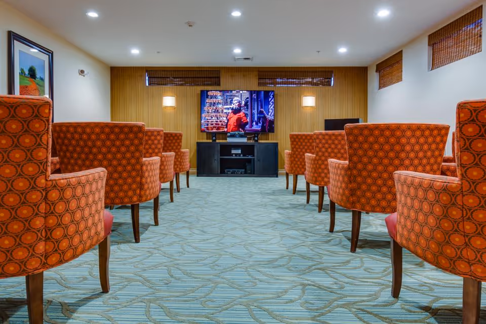 Rows of orange patterned armchairs in a carpeted media room facing a wall-mounted television.