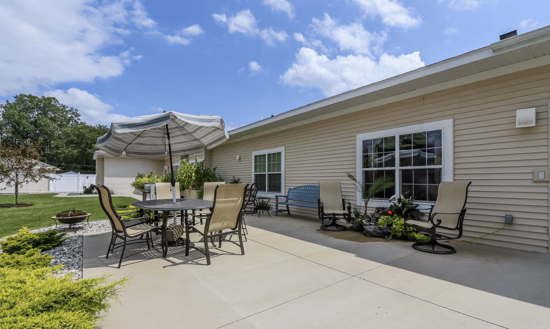 Outdoor patio area at Arbor Grove Assisted Living & Memory Care with a round table, four chairs, and a large striped umbrella. There are additional chairs and a bench along the building wall, with some potted plants and landscaping around the concrete patio. The sky is partly cloudy.