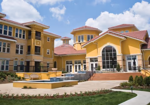 Exterior view of a large, yellow senior living facility building with multiple windows, red-tiled roofs, and a central entrance with steps leading up. There is a paved courtyard area with benches and landscaped greenery in front of the building under a partly cloudy blue sky.