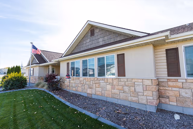 Single-story building exterior with a stone-and-siding facade, a row of windows, an American flag, and a manicured lawn.