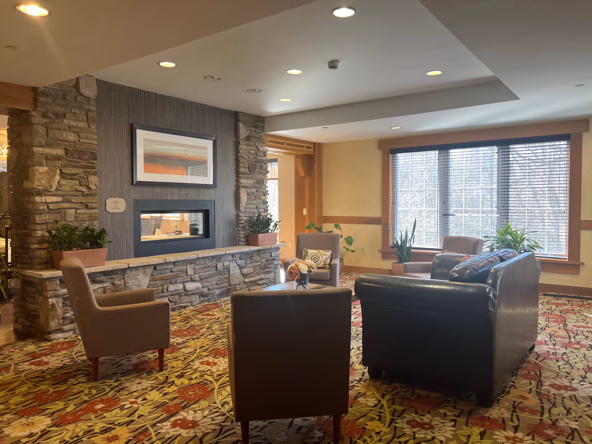 A cozy living room area with a stone fireplace, a framed abstract painting above it, and several chairs and a black leather sofa arranged around a small coffee table with a flower vase. Large windows with blinds allow natural light to fill the room, and there are several potted plants near the windows and fireplace. The carpet has a floral pattern with red, yellow, and green colors.