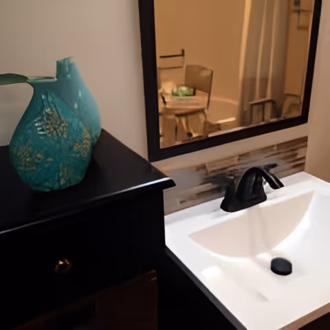 A bathroom sink with a black faucet and a white basin. Above the sink is a large mirror reflecting a small table and chair in the background. To the left of the sink is a black cabinet with a decorative turquoise vase on top.