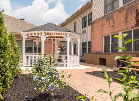Outdoor courtyard area with a white gazebo, flowering plants, a wooden bench, and a building with windows in the background under a partly cloudy sky.