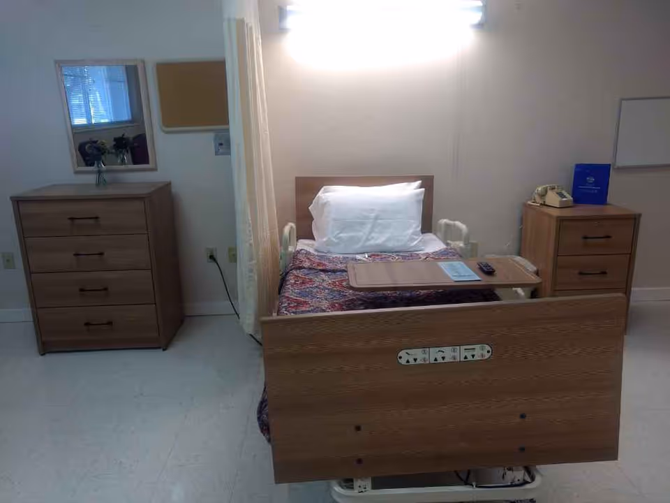 A single adjustable hospital-style bed with a tray table, pillow and patterned blanket flanked by wooden dressers in a plain patient bedroom.