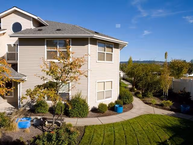 Two-story beige senior living building with landscaped lawn and curved concrete walkway.