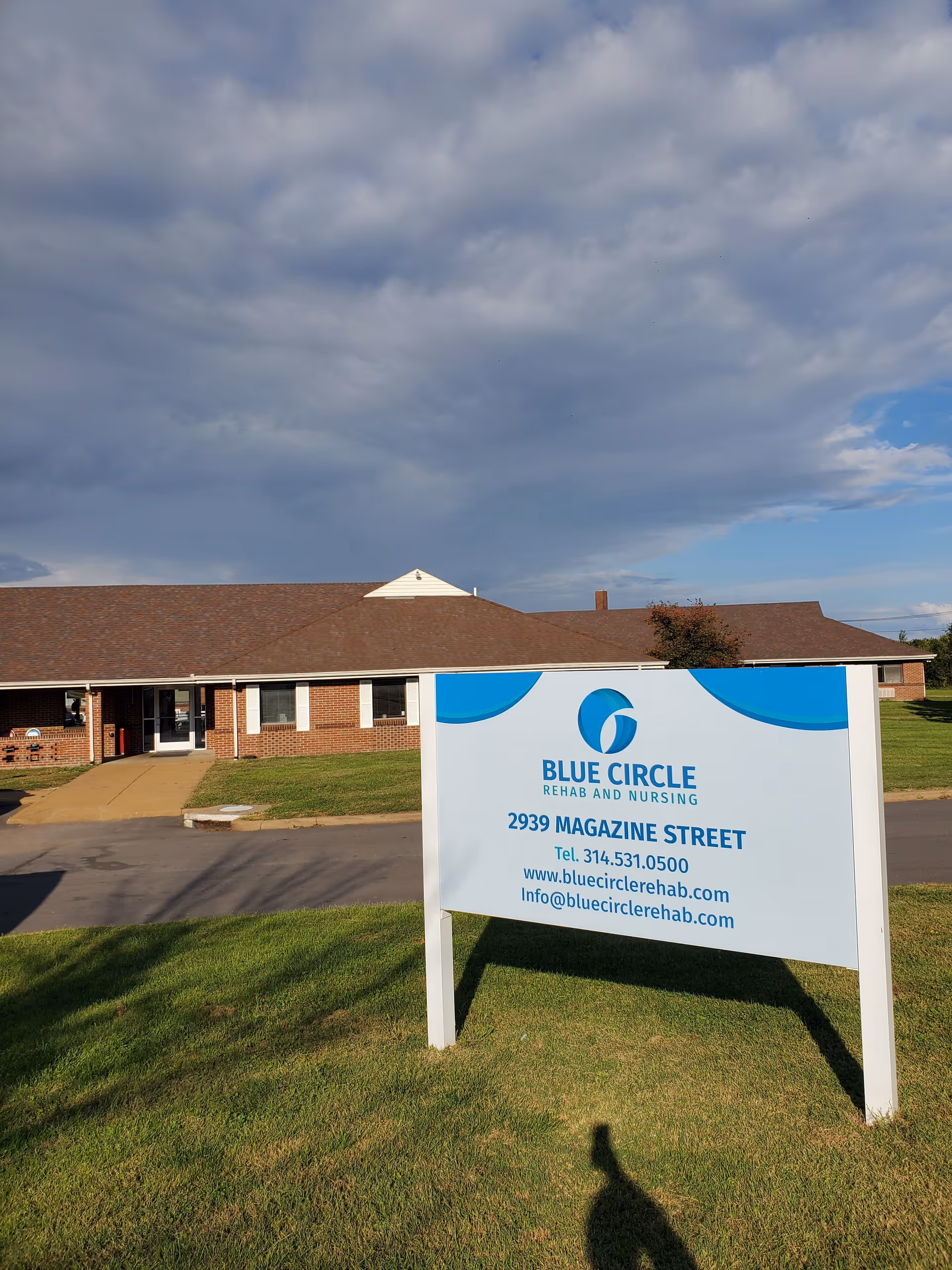 Exterior view of Blue Circle Rehab and Nursing facility with a large sign in the foreground displaying the facility's name, address, phone number, website, and email. The building is a single-story brick structure with a brown roof, set against a partly cloudy sky.