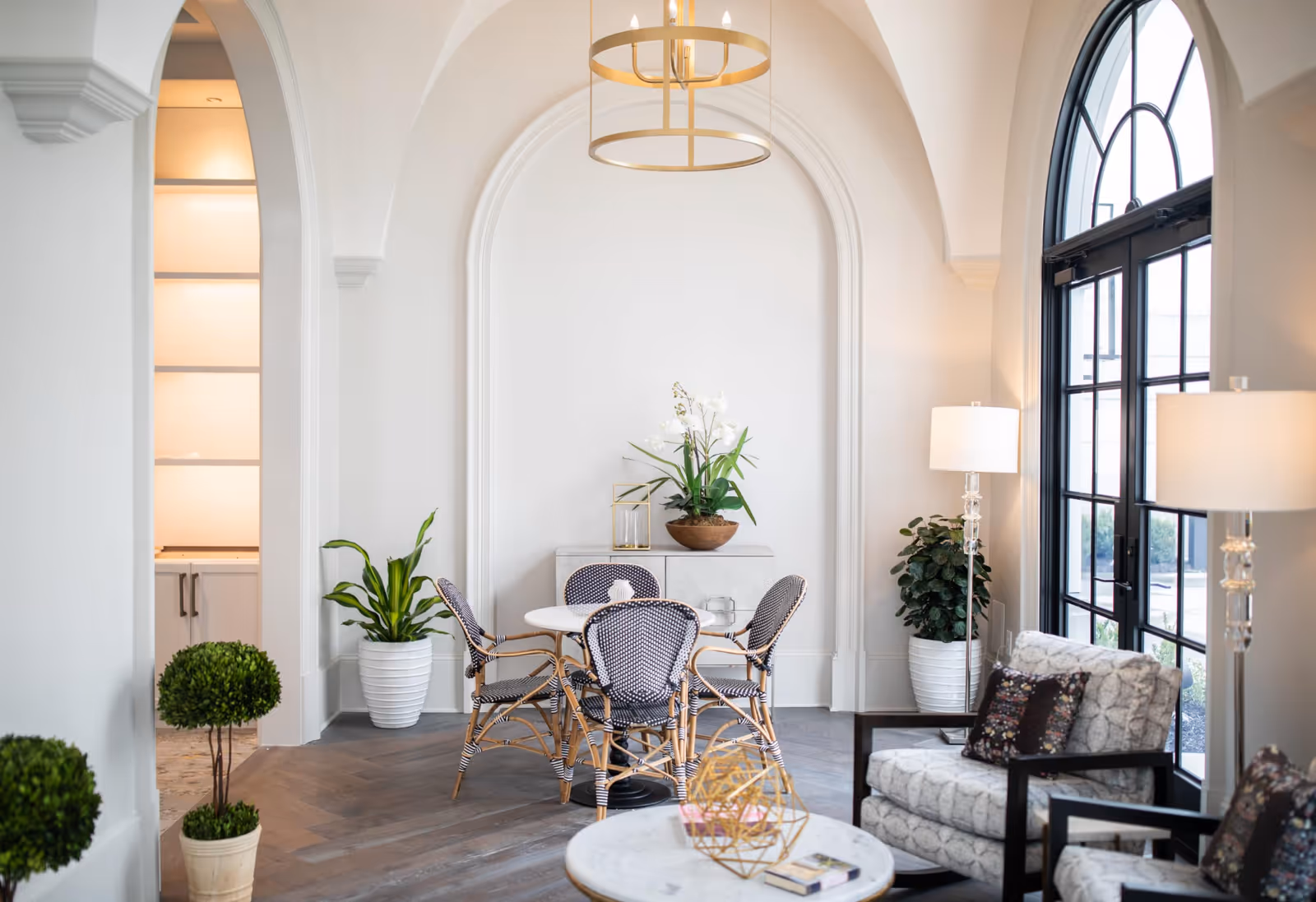 Bright and elegant interior space featuring a small round table with four woven chairs, potted plants, a sideboard with decorative items, two upholstered armchairs with patterned cushions, two floor lamps, and large arched windows allowing natural light to fill the room.