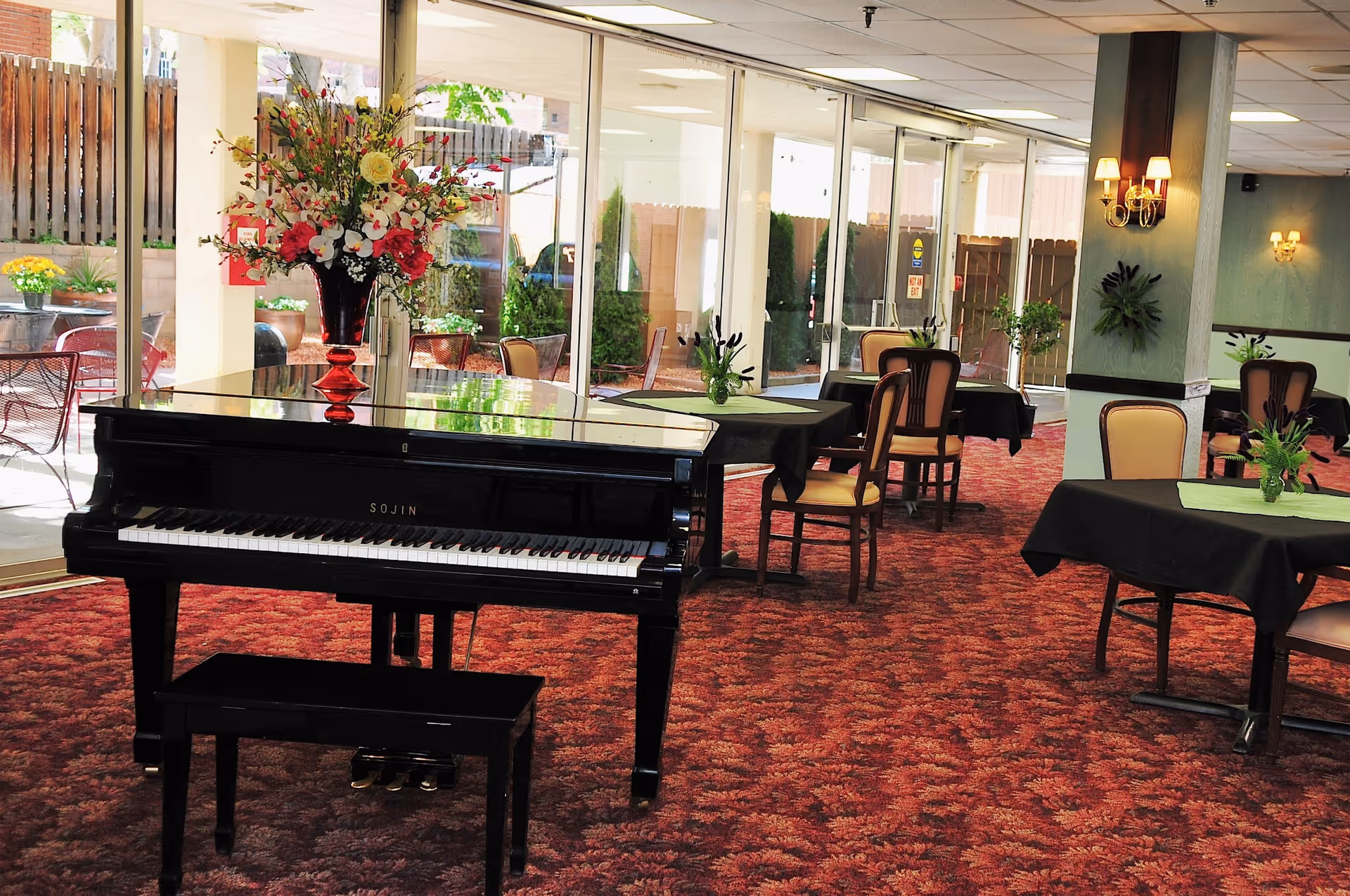 An indoor dining room with a black grand piano, a floral arrangement, and tables with chairs on red patterned carpet beside large glass windows.