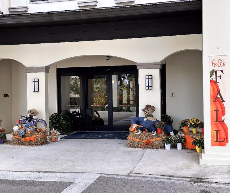 Entrance of Arbor Terrace Cooper City decorated for fall with two scarecrows sitting on hay bales, potted flowers, and a vertical banner that reads 'hello FALL' with pumpkin illustrations.