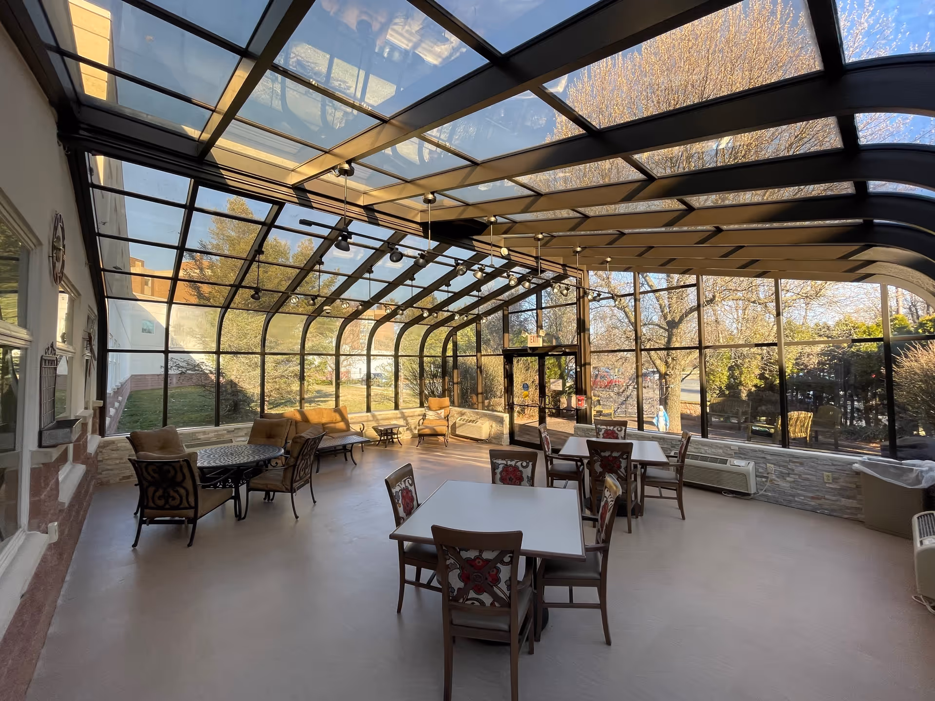 A sunroom with a glass ceiling and walls, furnished with several tables and chairs. The room has cushioned armchairs and a sofa along the left side, with a view of trees and greenery outside. The space is well-lit with natural light and has ceiling fans and track lighting.