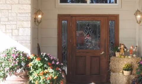 Front entrance of a building with a wooden door featuring decorative glass panels. The entrance is flanked by two wall-mounted lantern-style lights. There are potted plants with colorful flowers on the left side and a stack of hay bales decorated with small scarecrow figures and a potted plant on the right side.