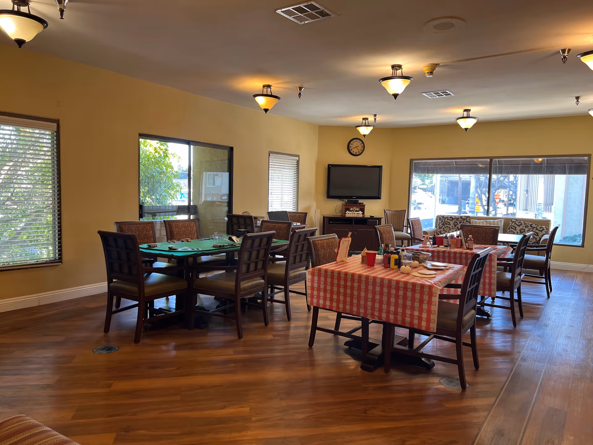 Communal dining room with multiple tables and chairs, one table covered with a red-checkered cloth, a wall-mounted TV and large windows.