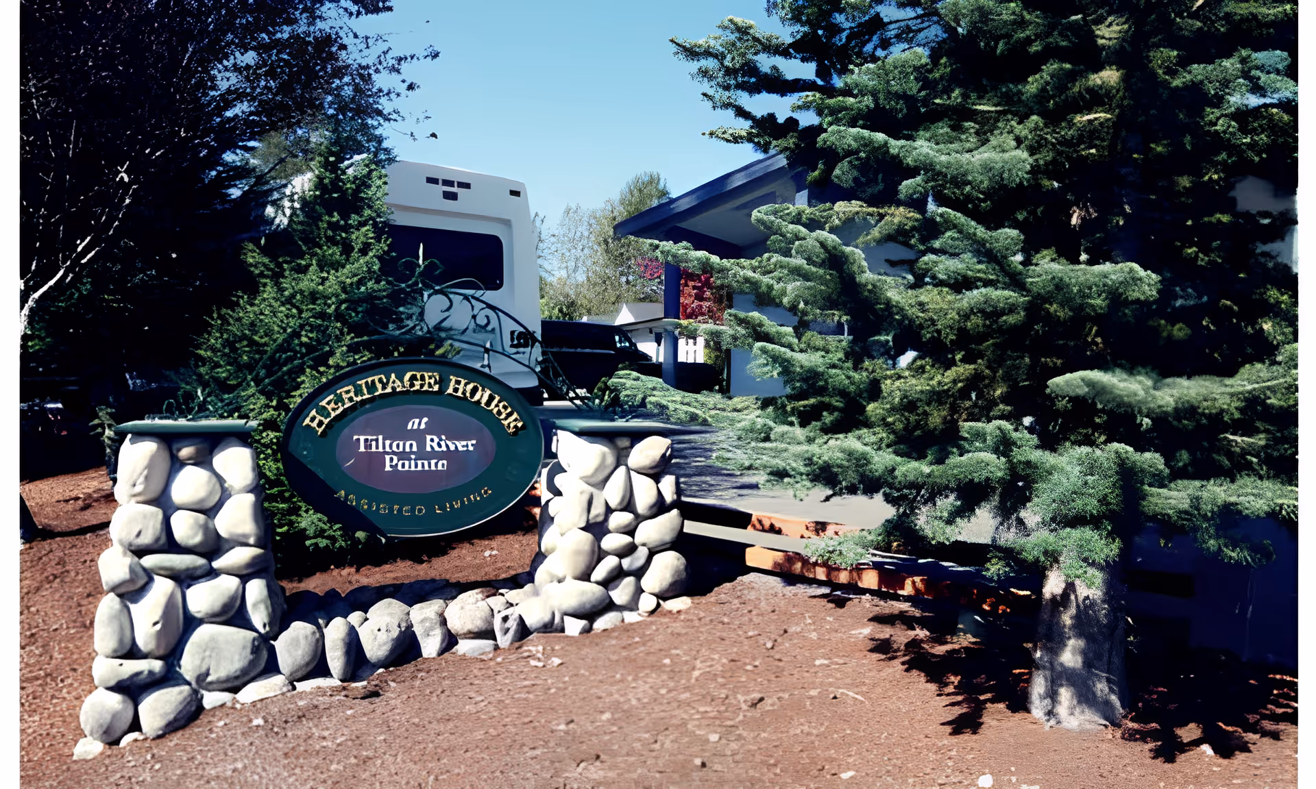 Stone sign for 'Heritage House' at the landscaped front of an assisted living building with trees and a driveway.