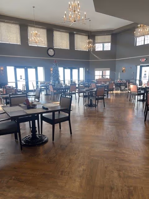 Spacious dining room with multiple tables and chairs arranged neatly on a wooden floor. Large windows and glass doors allow natural light to fill the room. A decorated Christmas tree is visible near the windows. Elegant chandeliers hang from the ceiling, and a clock is mounted on the wall above the windows.