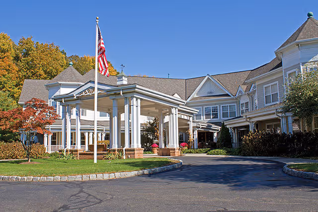Exterior view of a senior living facility building with a covered entrance, an American flag on a flagpole, and landscaped greenery including trees and bushes under a clear blue sky.