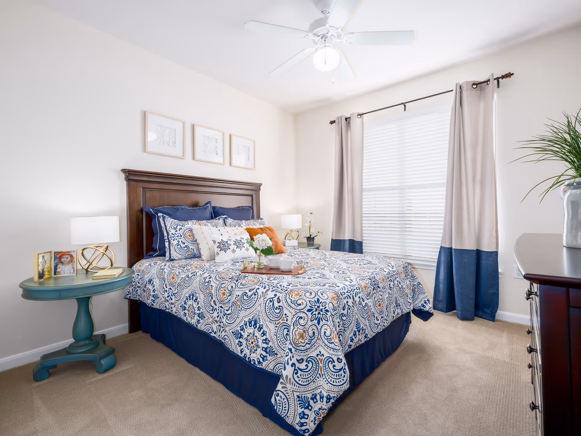 A bright and neatly arranged bedroom with a large bed featuring a blue and white patterned bedspread and multiple pillows. The room has beige carpet, a ceiling fan with light, a window with beige and blue curtains, a green round side table with a lamp, photo frames, and decorative items. A dark wooden dresser with a plant on top is visible on the right side.