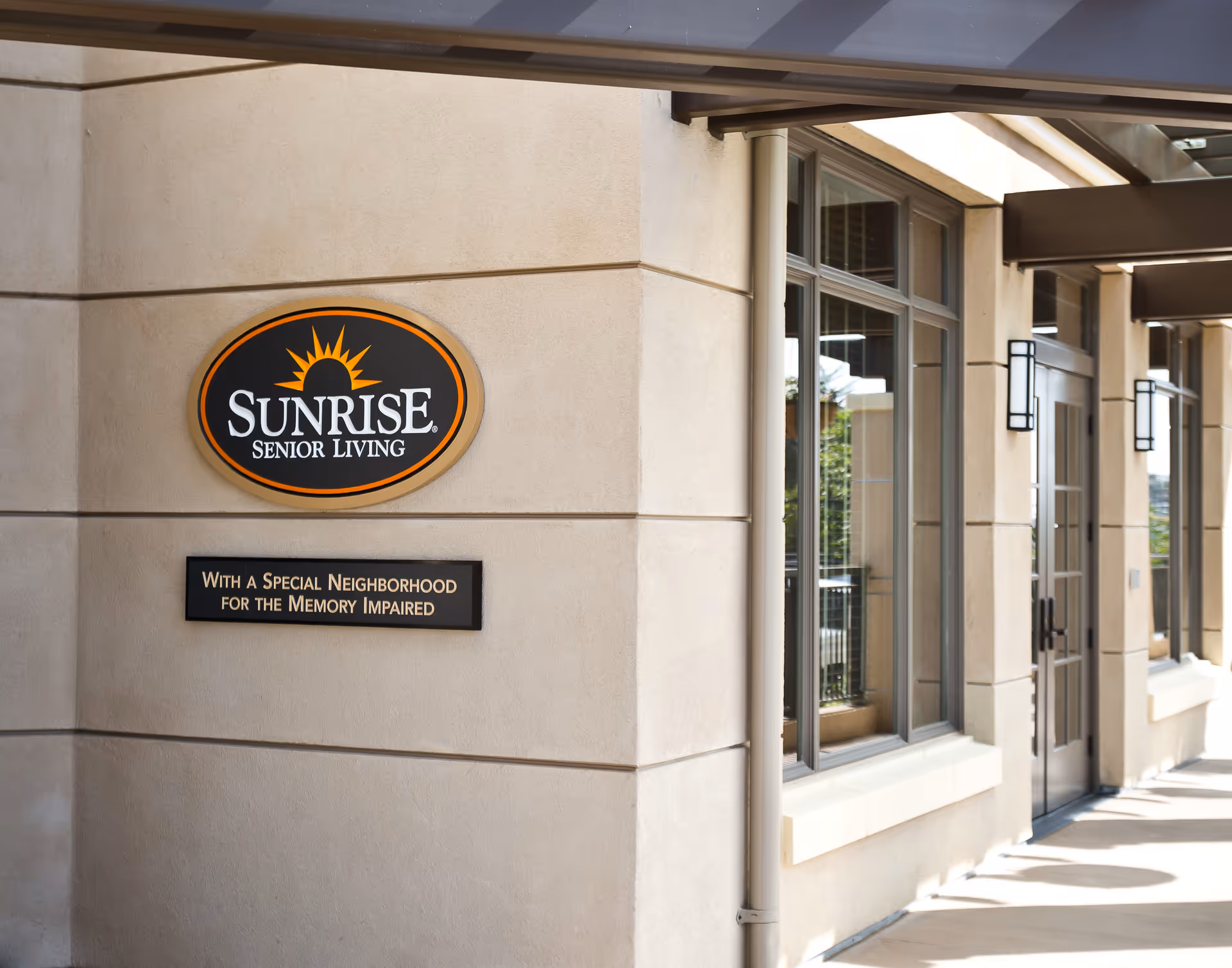 Exterior view of a senior living facility with a beige stucco wall featuring a Sunrise Senior Living sign and a smaller sign below that reads 'With a Special Neighborhood for the Memory Impaired'. There are large windows and a glass door along the building's facade under a covered walkway.