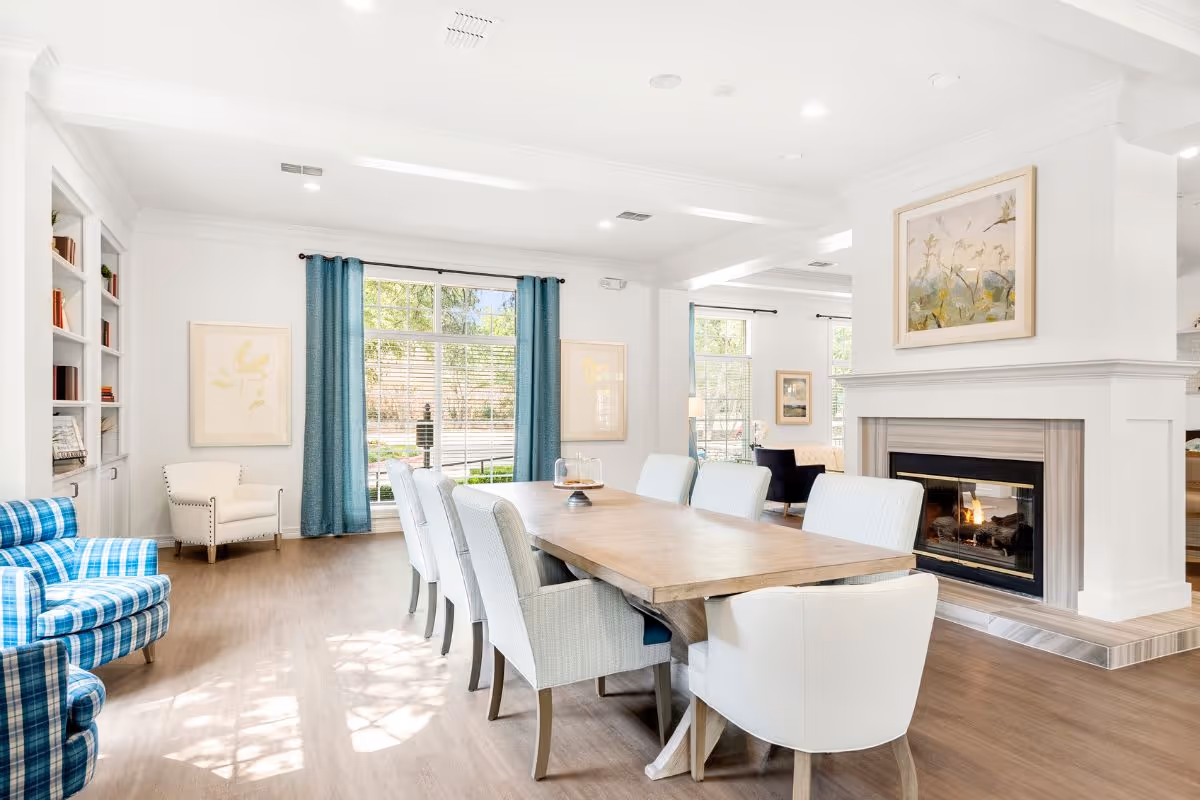Bright common dining area with a long wooden table surrounded by upholstered chairs, a fireplace, large windows with blue curtains, and seating areas.