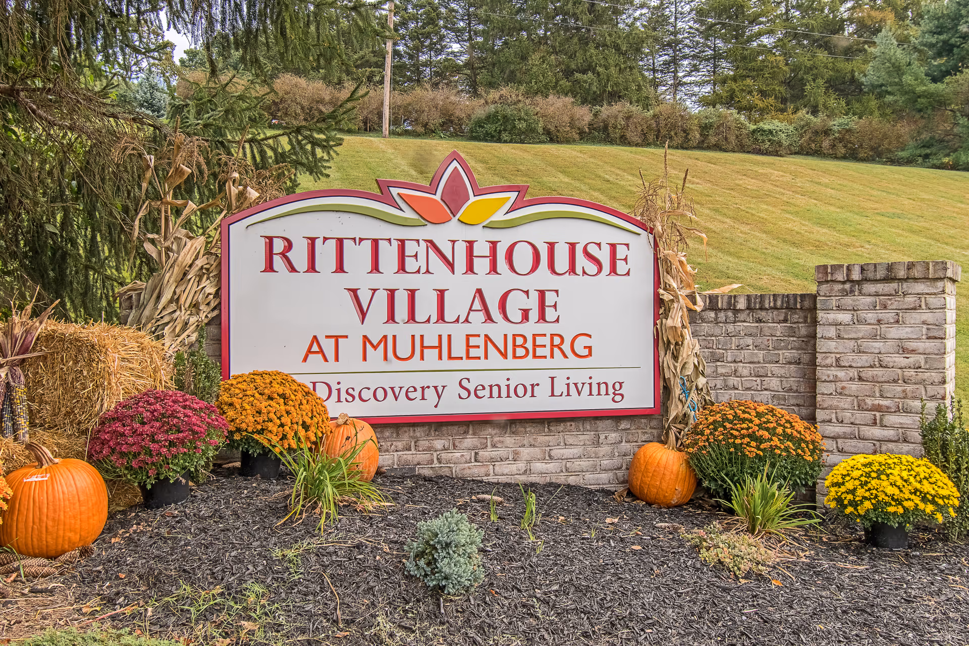 Entrance sign for Rittenhouse Village at Muhlenberg Discovery Senior Living decorated with pumpkins and fall mums in a landscaped brick planter against a grassy hillside.