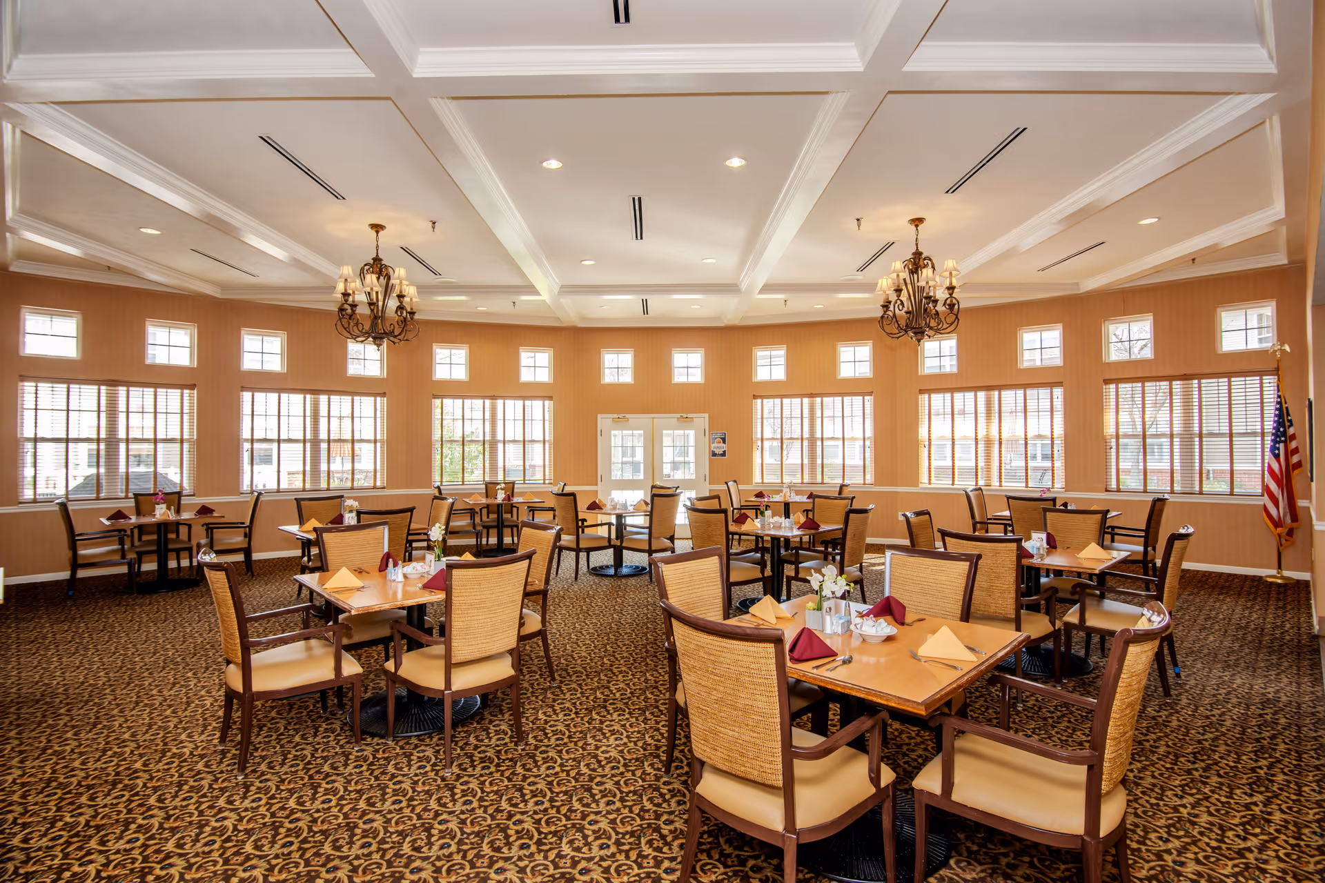 A spacious dining room with multiple wooden tables and chairs arranged neatly. Each table is set with folded napkins, utensils, and small floral centerpieces. The room has large windows with blinds allowing natural light to fill the space, ornate chandeliers hanging from a coffered ceiling, and an American flag in the corner.