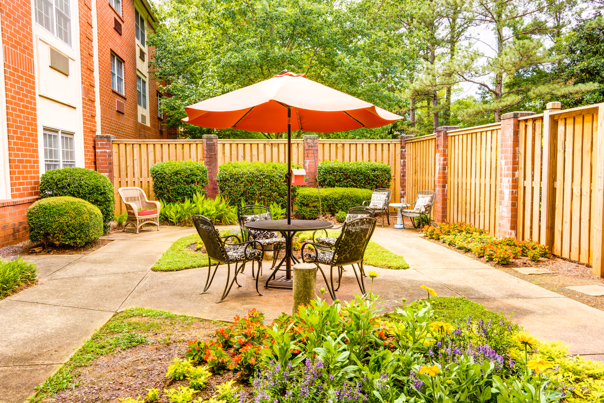 Outdoor patio area with a round table and four metal chairs under an orange umbrella. The patio is surrounded by a wooden fence and brick pillars, with neatly trimmed bushes and colorful flowers in the foreground. A brick building is visible on the left side.