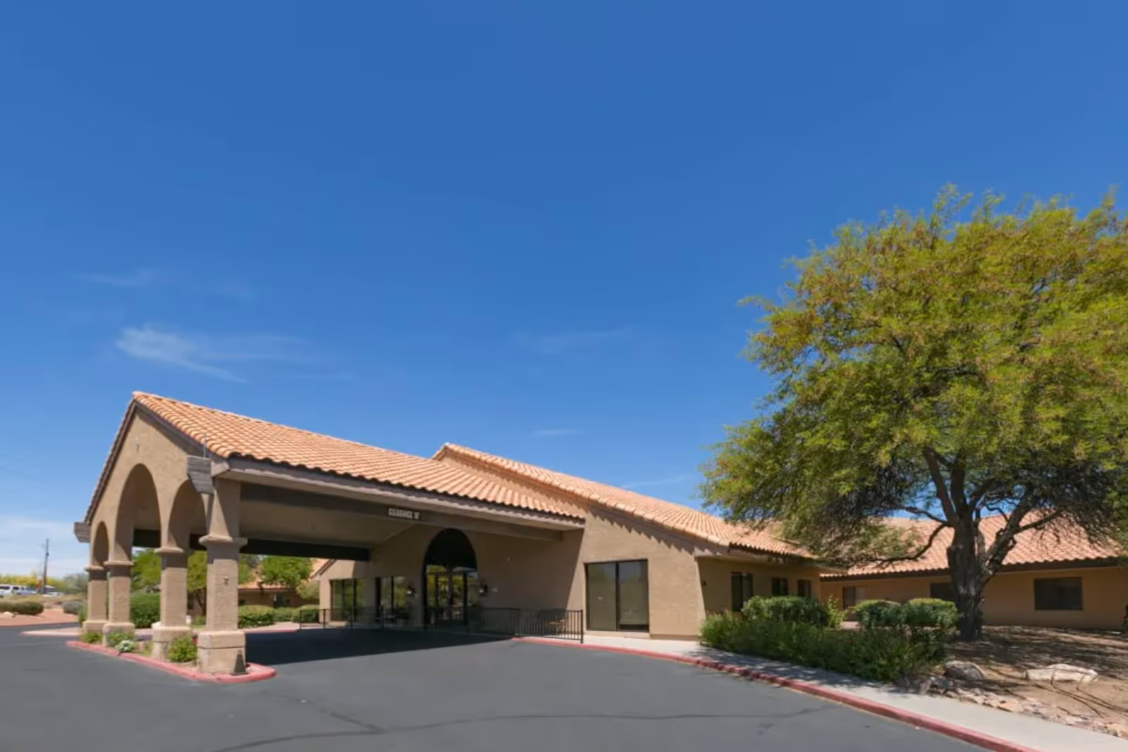 Front entrance of a single-story care facility with a tiled roof, covered porte-cochere, and landscaping including a large tree.