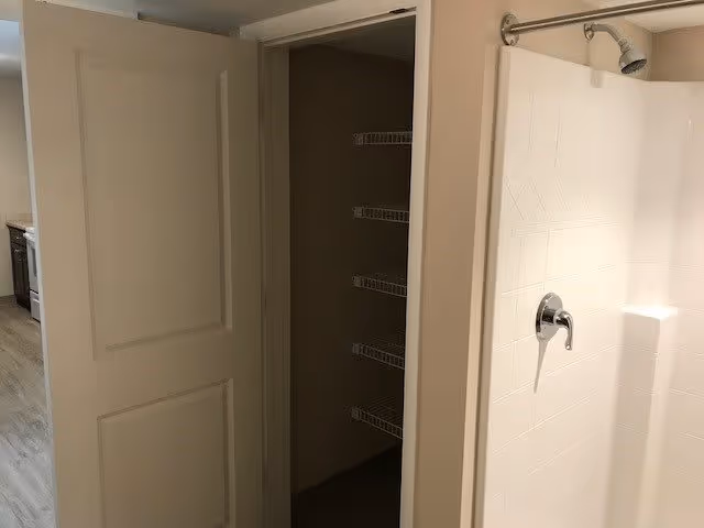 Interior view showing a partially open door revealing a closet with wire shelves, next to a white tiled shower area with a chrome showerhead and handle.