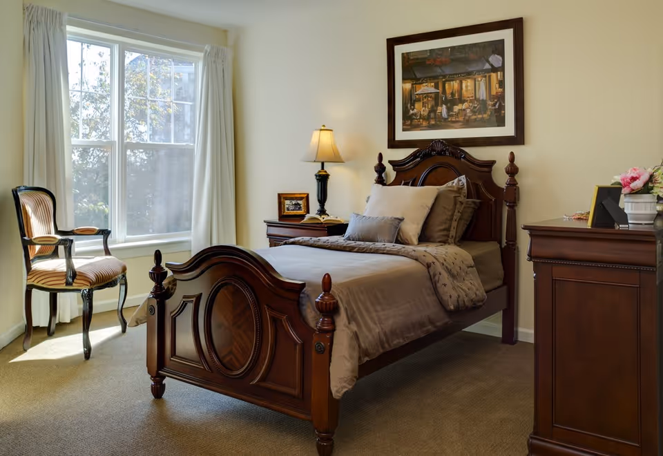Sunlit bedroom with an ornate wooden bed, matching nightstand and dresser, a chair by the window, lamp, and framed artwork.