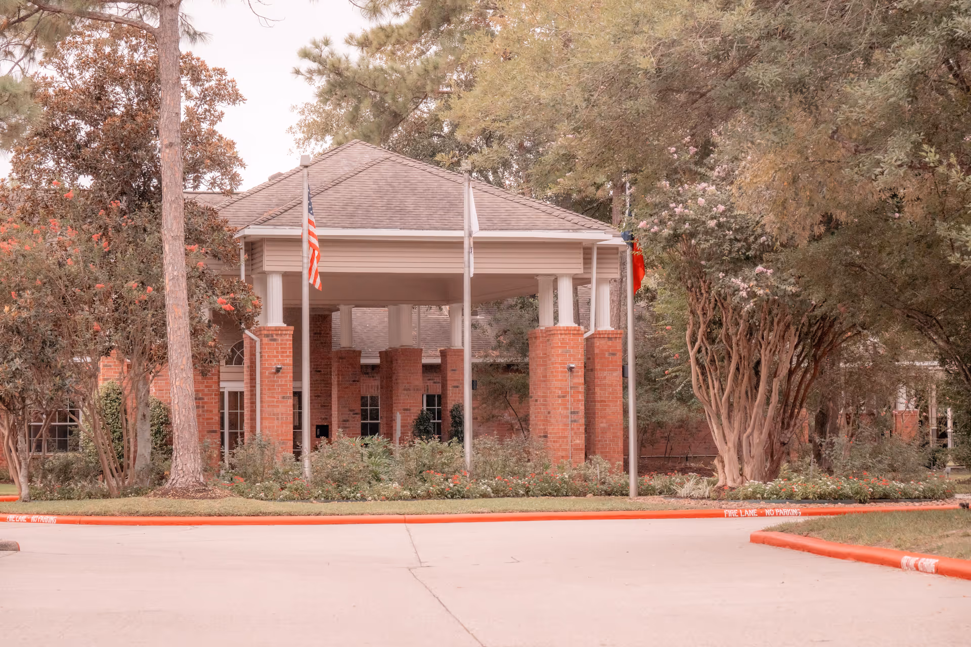 Front exterior view of a brick building with a covered entrance supported by white columns. There are three flagpoles with flags in front of the entrance, surrounded by trees and shrubs. The curb is painted red with 'FIRE LANE NO PARKING' written on it.