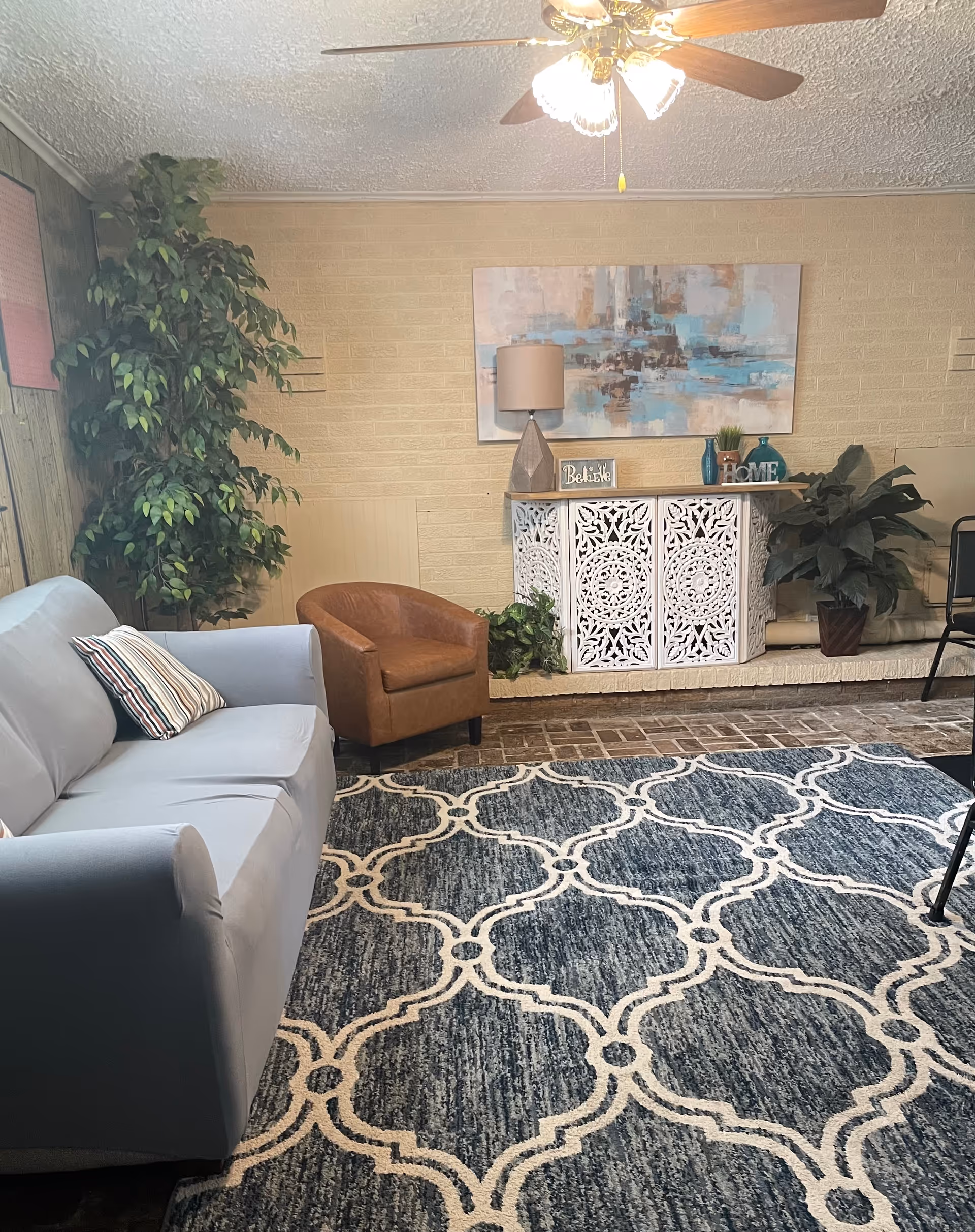 A cozy living room with a light blue sofa with a striped pillow, a brown leather armchair, a large patterned blue and white rug, a white decorative cabinet with intricate designs, a table lamp, and two potted plants. A ceiling fan with lights is visible above, and a large abstract painting hangs on the beige brick wall behind the cabinet.