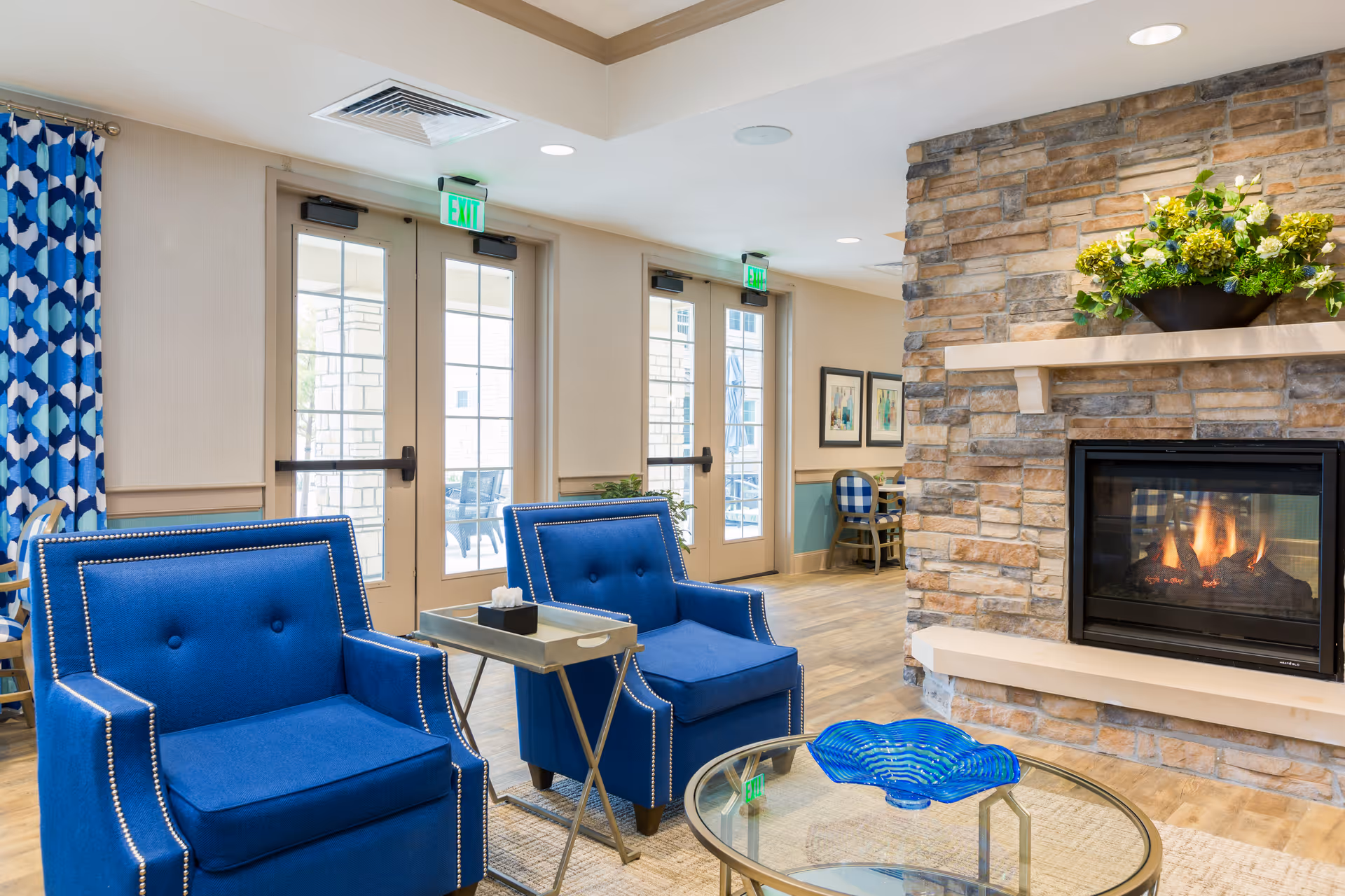 A cozy senior living facility common area with two blue armchairs featuring nailhead trim, a small side table with a tissue box, a glass coffee table with a blue decorative bowl, and a stone fireplace with a fire burning and a flower arrangement on the mantel. Behind the chairs are large glass doors leading to an outdoor patio area, and blue patterned curtains hang on the left side.