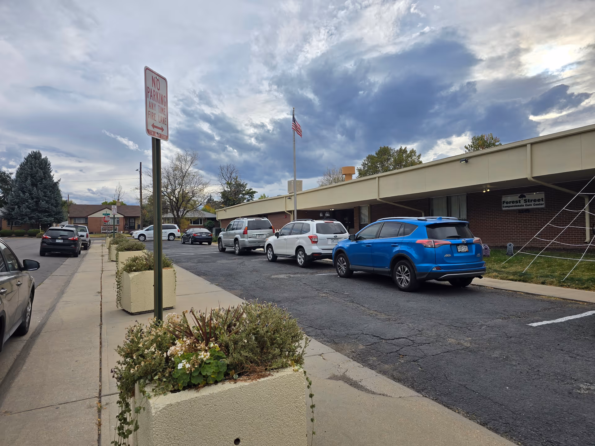 Exterior view of Forest Street Compassionate Care Center showing a single-story brick building with a flat roof, several parked cars along the driveway, an American flag on a flagpole, and a cloudy sky overhead. There are large planters with greenery along the sidewalk and a 'No Parking Any Time Fire Lane' sign visible.